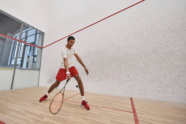joven afroamericano hombre en pantalones cortos deportivos rojos sosteniendo raqueta mientras juega squash, tiro de movimiento - Foto, Imagen