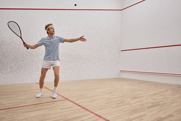 atlético pelirrojo hombre en ropa deportiva celebración de raqueta y jugar squash dentro de la cancha, tiro de movimiento - Foto, Imagen