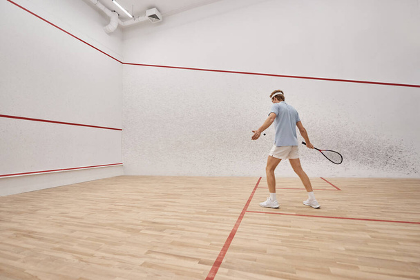 sporty and redhead man holding racquet while playing squash inside of court, motion shot - Fotó, kép