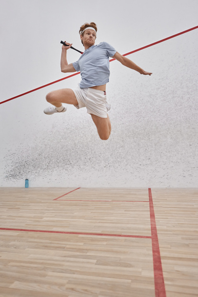 tiro en movimiento, deportista activo sosteniendo raqueta y saltando mientras juega squash dentro de la cancha - Foto, Imagen