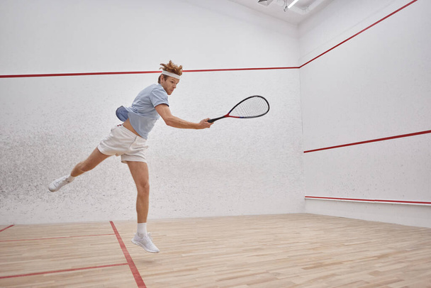 disparo de movimiento, jugador enérgico sosteniendo raqueta y saltando mientras juega squash dentro de la cancha - Foto, Imagen