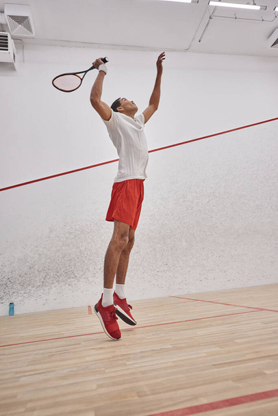 movimiento, enérgico hombre afroamericano sosteniendo raqueta mientras salta y juega squash, entrenamiento - Foto, imagen