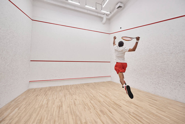 athletic african american player with racquet jumping and playing squash in court, back view - Foto, Imagem