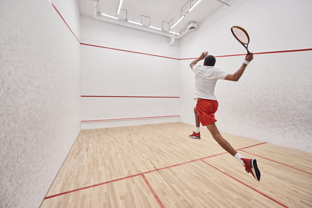 athletic african american player with racquet jumping while playing squash in court, back view - 写真・画像