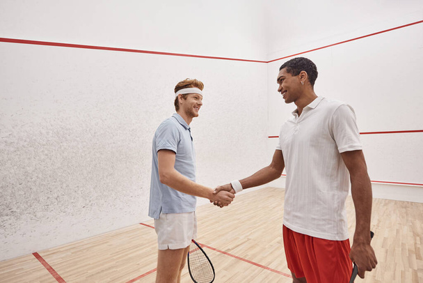 happy interracial sportsmen in active wear smiling and shaking hands while holding squash racquets - Photo, image