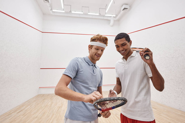joyful interracial sportsmen in active wear smiling and checking squash racquet inside of court - 写真・画像
