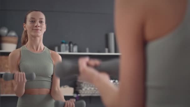 Közepes lövés az aktív fiatal kaukázusi nő élvezi a tükör, miközben csinál bicepsz göndör gyakorlat súlyzókkal a fedett edzőteremben edzés - Felvétel, videó