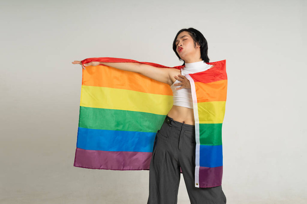 Asyalı eşcinsel bir adamın elinde gökkuşağı bayrağı tutarken beyaz bir arkaplanda güvenle poz vermesi. - Fotoğraf, Görsel