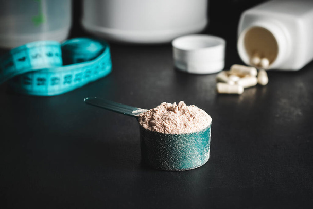 Chocolate whey protein powder in measuring spoon, white capsules of amino acids, vitamins and creatine, measuring tape, plastic shaker on dark background. bodybuilding food supplements. - Photo, Image