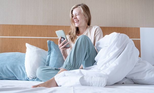 Γυναίκα χρησιμοποιώντας smartphone και γελώντας κάθεται στο κρεβάτι στο δωμάτιο του ξενοδοχείου - Φωτογραφία, εικόνα