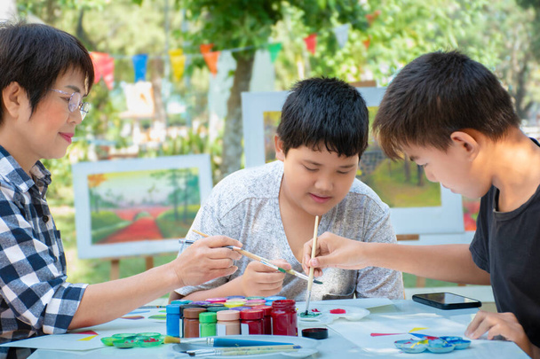 Aprender a usar el color del póster para dibujar varios dibujos de niños asiáticos en una mesa fuera del aula con su maestra de arte, criar a adolescentes y adultos ayuda a los niños a diseñar proyectos escolares. - Foto, imagen