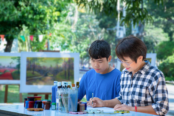 Aprender a usar el color del póster para dibujar varios dibujos de niños asiáticos en una mesa fuera del aula con su maestra de arte, criar a adolescentes y adultos ayuda a los niños a diseñar proyectos escolares. - Foto, imagen