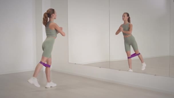 Νεαρή fit Καυκάσια γυναίκα στέκεται μπροστά από το full length καθρέφτη στον τοίχο σε φωτεινό ευρύχωρο γυμναστήριο που κάνει άσκηση απαγωγής ποδιών με ελαστική ζώνη αντίστασης - Πλάνα, βίντεο