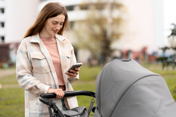 Jeune mère souriante utilisant un téléphone portable, marchant avec son nouveau-né en poussette. Belle femme en plein air dans le parc avec enfant, vérifier le courrier, achats en ligne - Photo, image