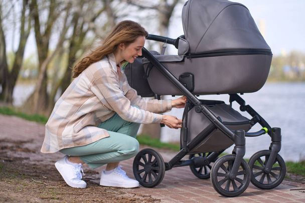 Junge Mutter, schöne kaukasische rothaarige Frau in lässiger Kleidung, die den Kofferraum im Kinderwagen während eines Spaziergangs mit ihrem neugeborenen Kind im Park am Fluss öffnet. - Foto, Bild