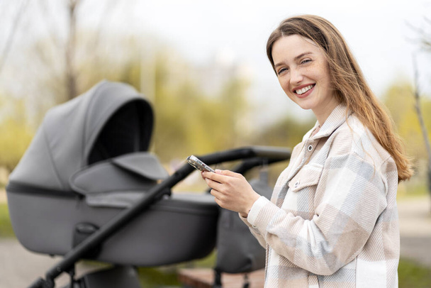 Smiling beautiful woman using smartphone outdoors, walking with stroller in park. Positive young mother looking at camera, doing online shopping - Photo, Image