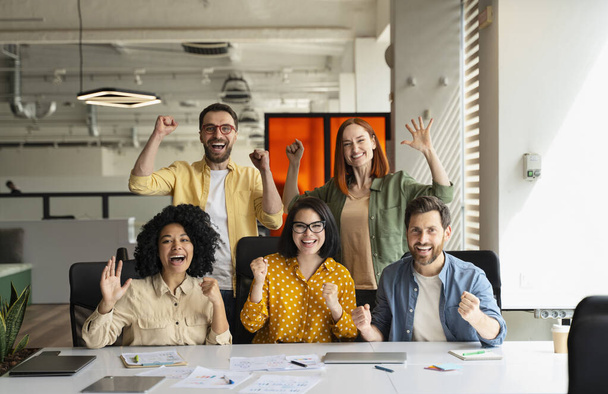 Успешная команда коллег по бизнесу, сжимая кулаки от волнения, улыбаясь, глядя в камеру, сидя за столом с ноутбуком, графиками и диаграммами в современном корпоративном интерьере офиса - Фото, изображение
