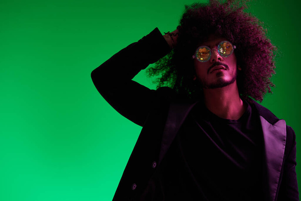 Retrato de moda de um homem com cabelo encaracolado em um fundo verde com óculos de sol, multinacional, luz rosa colorida, moderno, conceito moderno. Foto de alta qualidade - Foto, Imagem