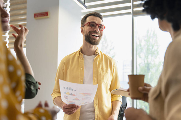 Красивый кавказский бородатый мужчина в стильных очках, держит в руках документ с диаграммами роста продаж, улыбается, разговаривая со своими коллегами, делится идеями и стратегиями развития бизнеса в современном офисе - Фото, изображение