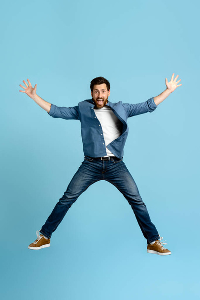 Full length verticaal studio portret van een blanke lachende emotionele knappe bebaarde man in de 40, springen hoog op en strek zijn handen asides, op geïsoleerde blauwe achtergrond - Foto, afbeelding