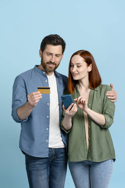 Mavi arka planda izole edilmiş plastik kredi kartı ve cep telefonu tutan beyaz mutlu bir çift. İnternetten alışveriş. İnternet bankacılığı. Nakitsiz ödeme. Faturaları ödüyorum. rezervasyon ve yemek siparişi - Fotoğraf, Görsel