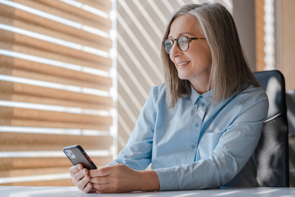 Femme d'affaires âgée souriante tenant un téléphone portable lisant un message texte, communication en ligne  - Photo, image