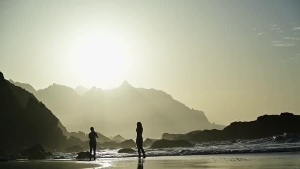 若いカップルは,テネリフェのスマートフォンバックライトを使用してビーチの日没で写真を撮ります - 映像、動画