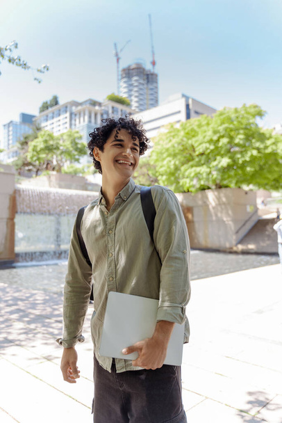 Egy mosolygós, jóképű latin férfi képe, aki laptoppal a kezében a város utcai kamerát nézi. Az online oktatás koncepciója, modern technológia a tanulásban - Fotó, kép