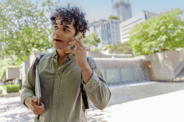 Πορτρέτο του χαμογελαστού όμορφου Ισπανόφωνου μαθητή να μιλάει στο κινητό, να απαντάει να κοιτάει αλλού στον αστικό δρόμο. Επιτυχημένες επιχειρήσεις - Φωτογραφία, εικόνα