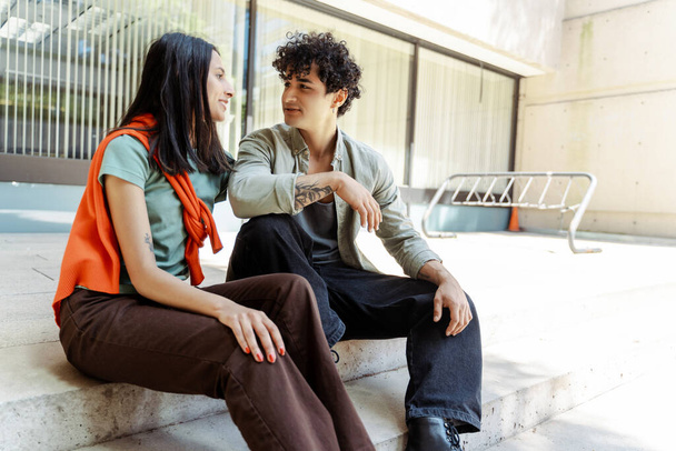 Cheerful friends sitting on stairs talking. Young students on street near university campus. Education concept - 写真・画像