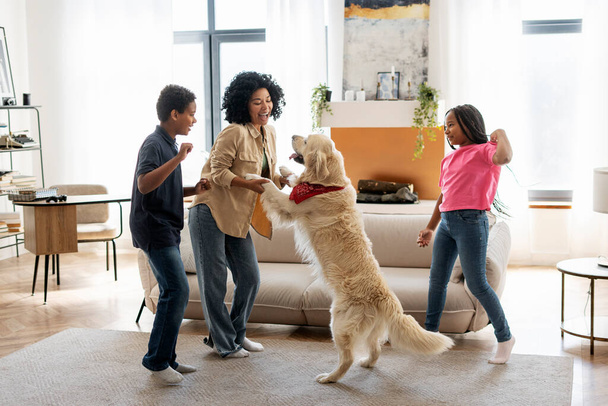Portrait of smiling beautiful African American family, mother, daughter and son playing dancing with golden retriever at home in living room. Children having fun. Happy childhood concept - Photo, Image