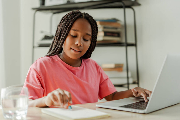 Belle fille afro-américaine sérieuse, écolière, assise à table, prenant des notes, utilisant un ordinateur portable, faisant ses devoirs. Écolière intelligente se préparant pour l'examen. Concept de retour à l'école - Photo, image