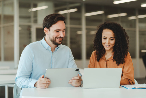 Портрет привлекательных улыбающихся бизнесменов, сидящих на рабочем месте, использующих ноутбук и цифровой планшет, планирующих проект в современном офисе. Технологическая концепция, успешный бизнес - Фото, изображение
