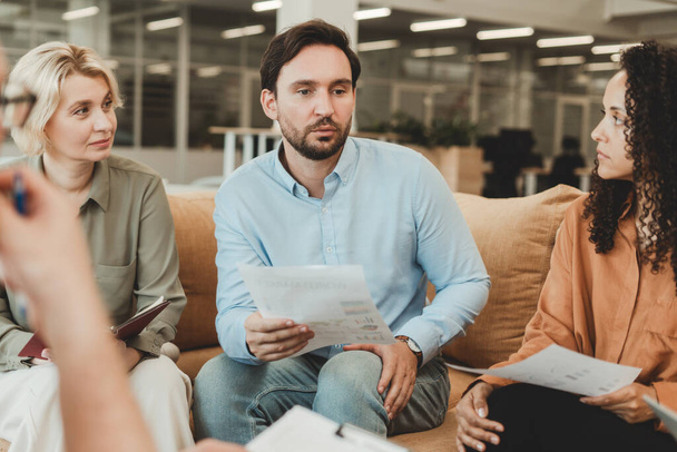 Confident bearded manager wearing casual clothes sitting on comfortable sofa in modern office and talking with his colleagues, holding document, working together, sharing ideas. Teamwork meeting - Photo, Image