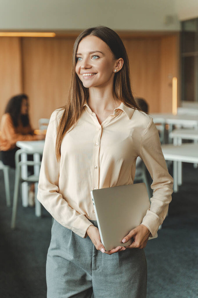 Atrakcyjna, dobrze wyglądająca młoda bizneswoman trzymająca laptopa pracująca w nowoczesnym biurze, odwracając wzrok. Uśmiechnięta bizneswoman, odnosząca sukcesy przedsiębiorca. Koncepcja przedsiębiorstwa - Zdjęcie, obraz
