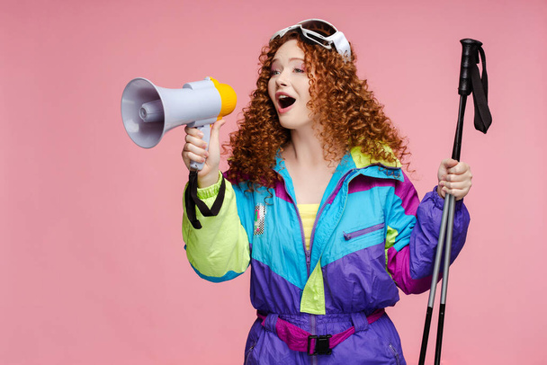 Portrait of positive curly skier woman wearing stylish ski overalls, goggles holding megaphone announcing black friday sale isolated on pink background. Shopping concept - 写真・画像