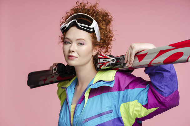 Portrait of beautiful curly redhaired woman, tourist wearing stylish vintage ski overalls, looking at camera, holding skis. Concept of winter travel, hobby - Photo, Image