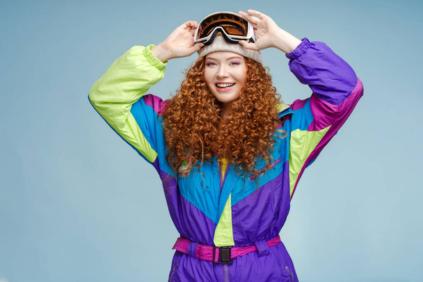 Portrait d'une jolie femme aux cheveux bouclés et souriante portant un chapeau et des lunettes de ski protectrices, des combinaisons de ski vintage posant isolées sur fond bleu. Concept d'hiver, voyage, publicité - Photo, image