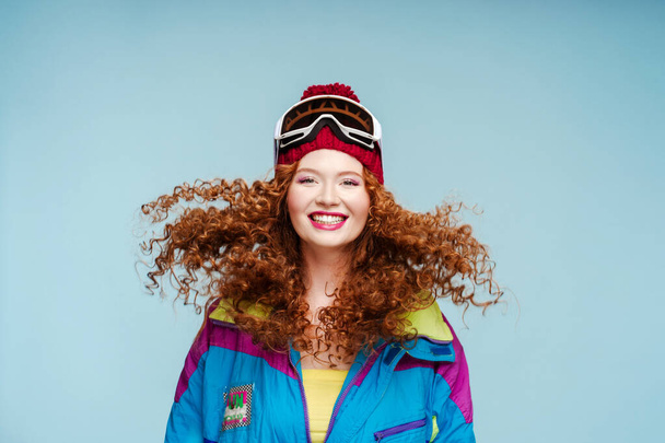 Belle femme bouclée rousse souriante portant des combinaisons de ski et des lunettes de saut, regardant la caméra isolée sur fond bleu. Concept vacances d'hiver - Photo, image