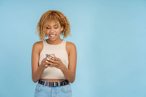 Krásná usmívající se Afroameričanka s rovnátka drží mobilní telefon komunikace on-line izolované na modrém pozadí, kopírovat prostor. Technologický koncept - Fotografie, Obrázek