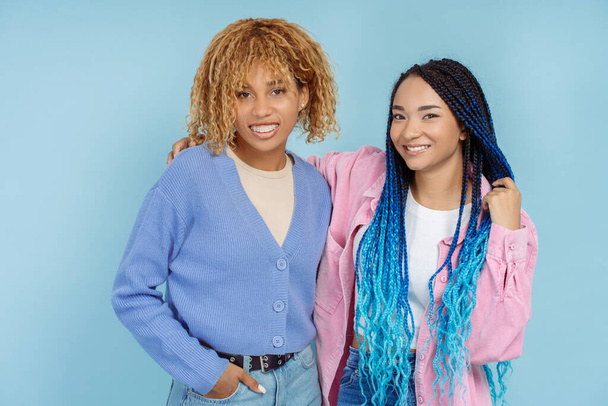Portrait smiling african american women in colorful casual clothes with braids looking at camera isolated on blue background. Positive female friends hugging. Concept of friendship - Photo, Image