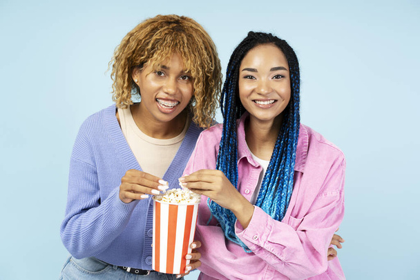 Young smiling African American women, best friends eating popcorn watching movie isolated on blue background - Foto, immagini
