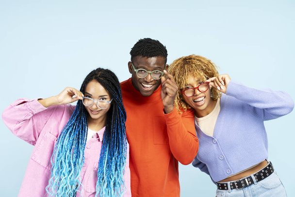 Group of smiling African American friends wearing colorful eyeglasses isolated on blue background. Happy fashion models with stylish hair looking at camera posing for pictures in studio - Photo, Image