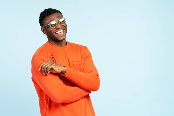 カジュアルなオレンジ色のTシャツを着ている魅力的なアフリカ系アメリカ人の男性の笑顔の肖像画,青い背景に隔離されたスタイリッシュな眼鏡. ショッピング,広告のコンセプト - 写真・画像