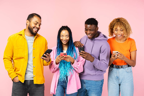 Grupa uśmiechnięta atrakcyjnych Afroamerykańskich przyjaciół trzymających smartfony za pomocą nowoczesnej aplikacji mobilnej zakupy online odizolowane na różowym tle. Stylowe kobiety i mężczyźni oglądają filmy na stronie internetowej. Technologia - Zdjęcie, obraz