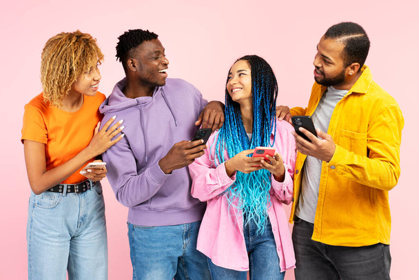 Grupa atrakcyjnych uśmiechniętych afro-amerykańskich przyjaciół trzymających telefony komórkowe, komunikujących się, rozmawiających na różowym tle. Technologia, koncepcja przyjaźni - Zdjęcie, obraz
