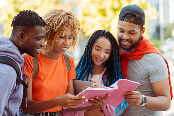 Группа улыбающихся молодых афроамериканских студентов, читающих книги, готовящихся к экзаменам, стоящих возле университетского городка на улице. Счастливые молодые люди встречаются на открытом воздухе - Фото, изображение