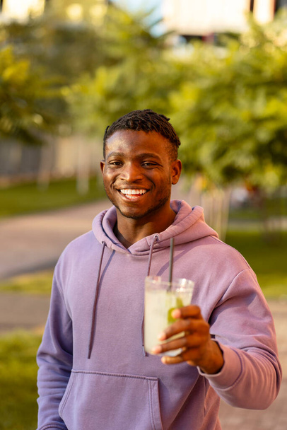 Przystojny, uśmiechnięty, stylowy Afroamerykanin, młody mężczyzna trzymający lemoniadę, pijący, patrzący w kamerę na ulicy, na zewnątrz - Zdjęcie, obraz