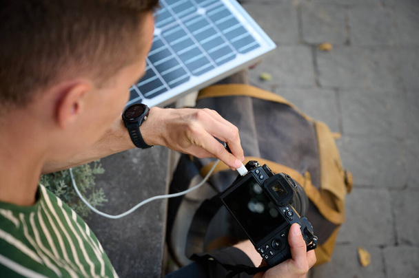 Utazó fotós, aki napelemet használ, hogy feltöltse a kameráját. Egy fiatal utazó várja, hogy a kamerája feltöltődjön. Pozitív férfi pihentető hosszú séta után. - Fotó, kép
