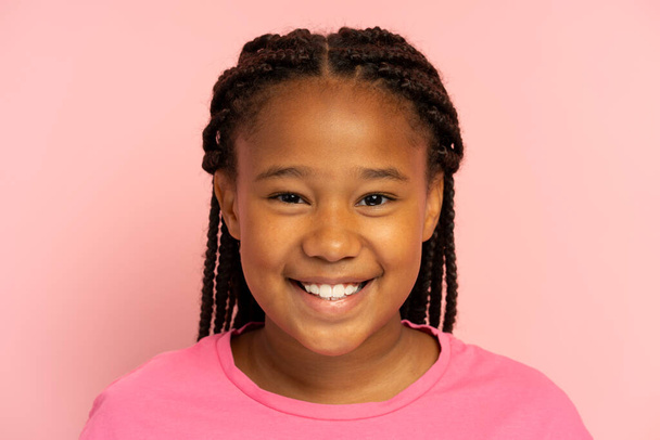 Portrait of cute smiling African American girl wearing stylish hairstyle, dreadlocks, pigtails wearing casual pink t shirt standing isolated on pink background, looking at camera, closeup - 写真・画像